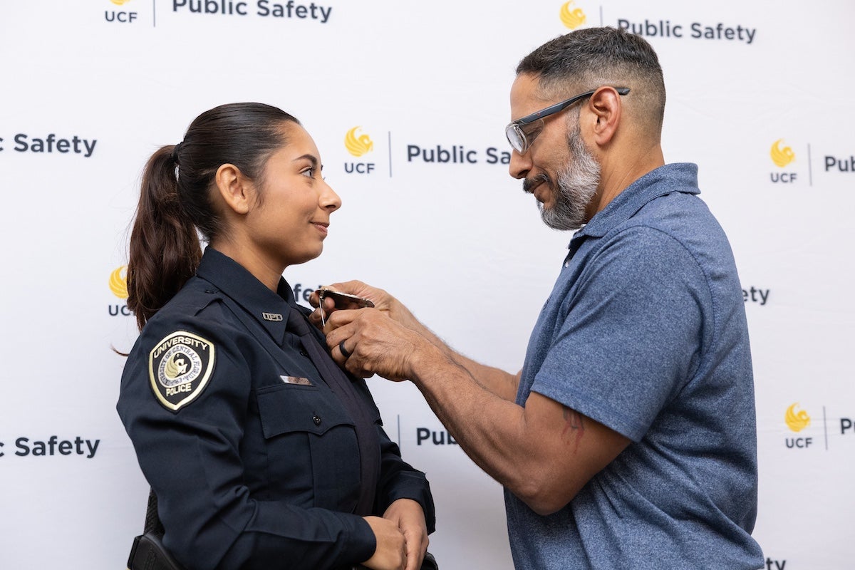A new UCF Police Department officer stands in uniform as her father pins her badge during a swearing-in ceremony.