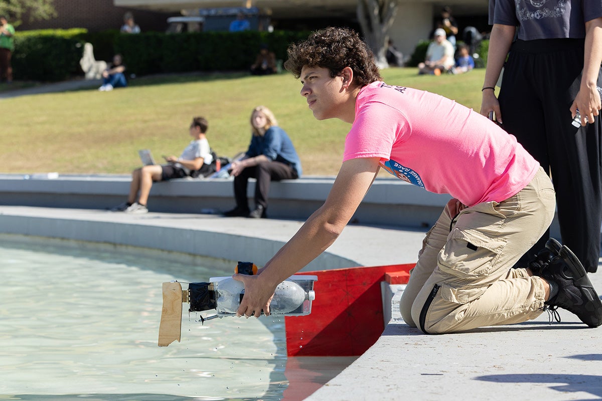 Male student with curly brown hair in pink shirt and khaki pants kneels at edge of pond, holding a small constructed boat above clear water.