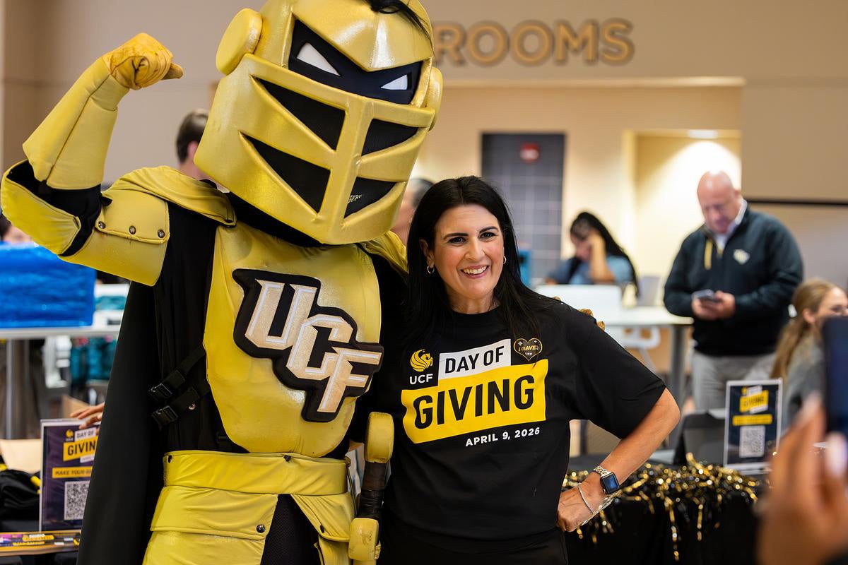 Brunette woman wearing black UCF Day of Giving T Shirt poses with hand on hip next to Knightro flexing in the Student Union