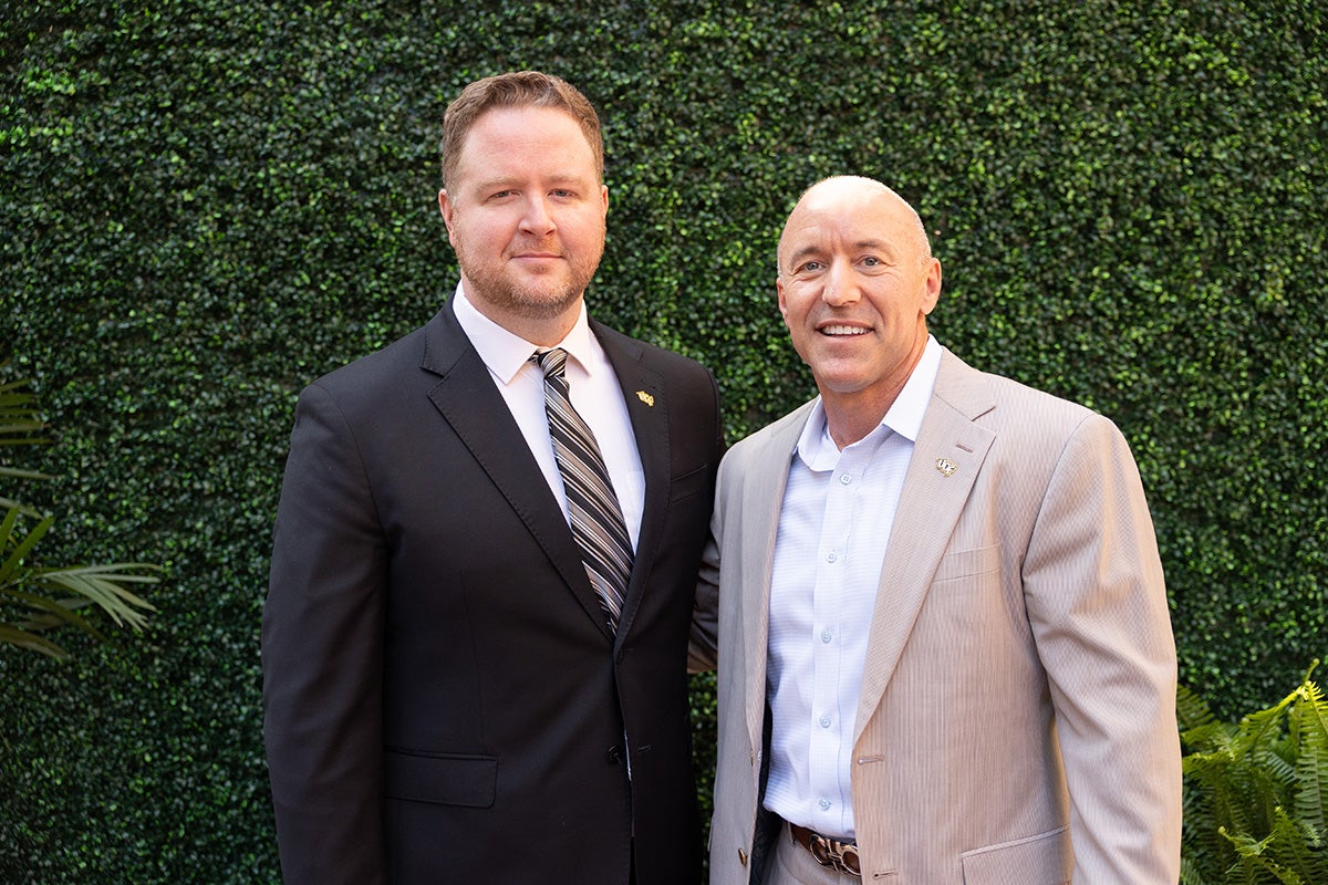 Alex Hubenthal in dark suit, white dress shirt and tie, stands to the right of Barry Miller in beige suit jacket and white shirt in front of grass wall