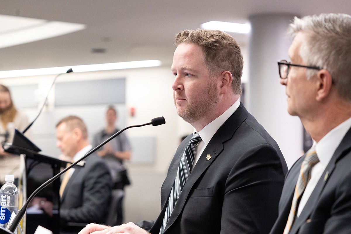 Bearded man in dark suit jacket, white dress shirt and tie sits at table with microphone as gray hair man with glasses observes from his left.