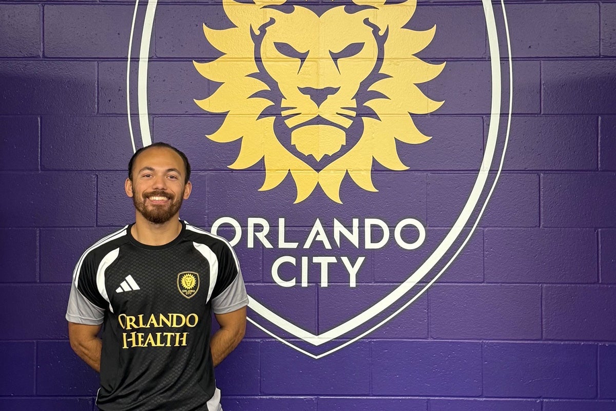 A man wearing an Orlando City soccer training jersey stands in front of a purple Orlando City logo wall.
