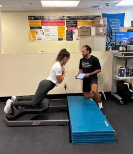 A researcher kneels beside a fellow researcher performing a hamstring strength test on specialized equipment in the Cognition, Neuroplasticity and Sarcopenia Lab at UCF.