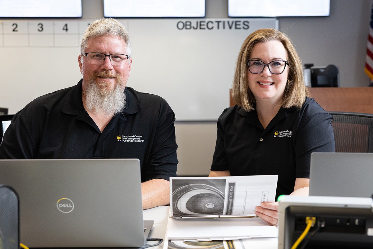 Short haired woman with glasses sits to the left of man with gray hair and beard, both wearing black polo shirts, with binders of paper and open laptop on desk in front of them and whiteboard behind them with "Objectives" in black letters at top of the board.