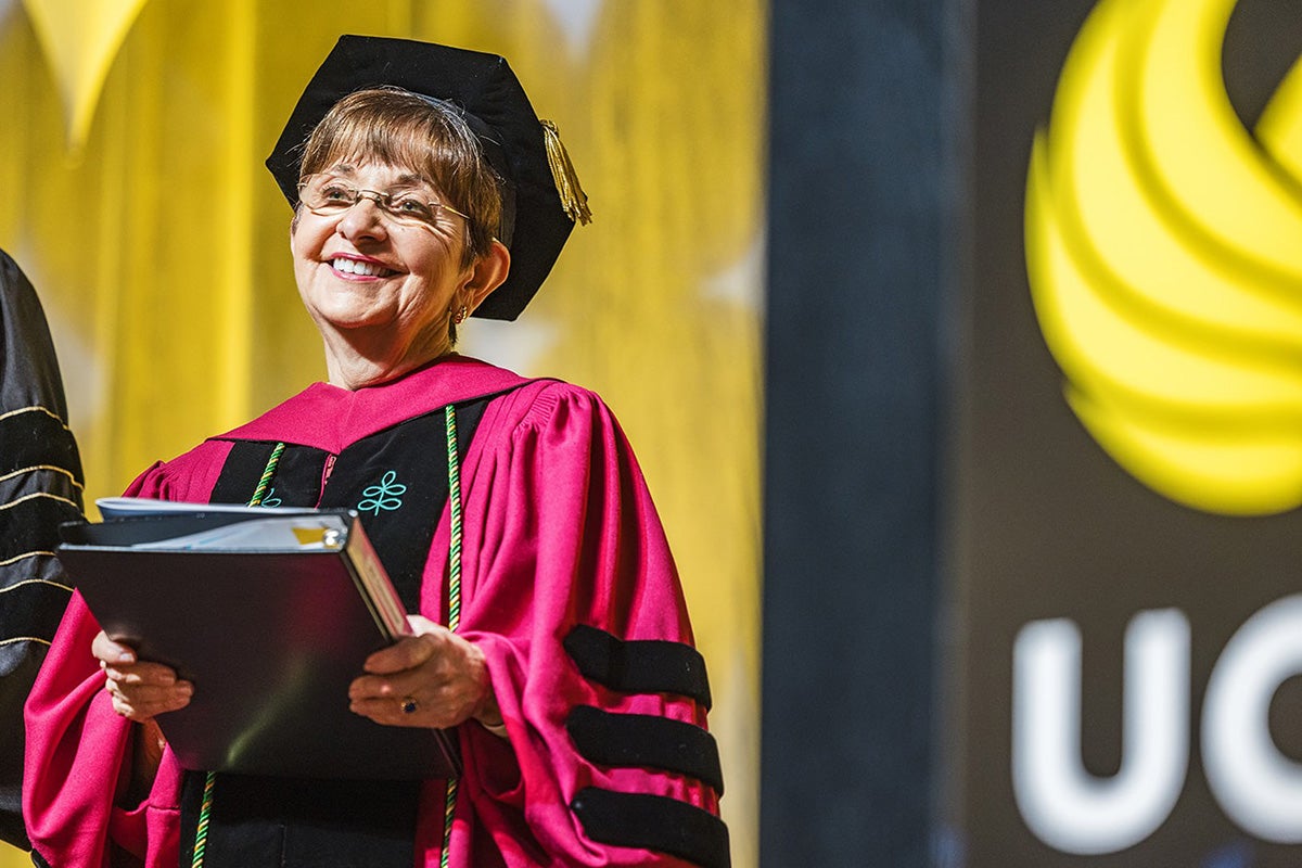 Deborah German in pinkish-red graduation gown and black cap smiles on stage with UCF logo behind her