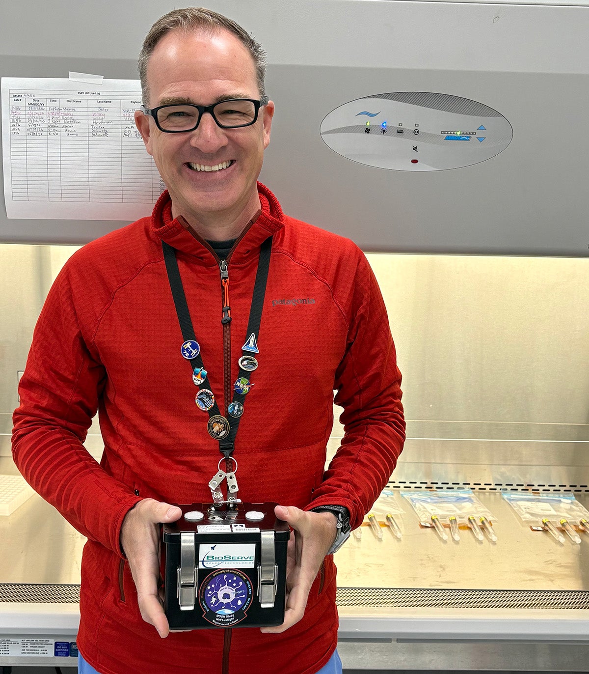 Man wearing glasses and red Patagonia pullover stands holding black and metal cube in his hands in front of gray lab equipment