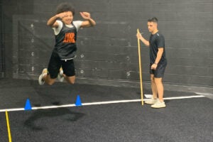 A student-athlete jumps over cones during a training drill while UCF alum Jeremy Wydra observes and holds a measuring stick in a gym setting.
