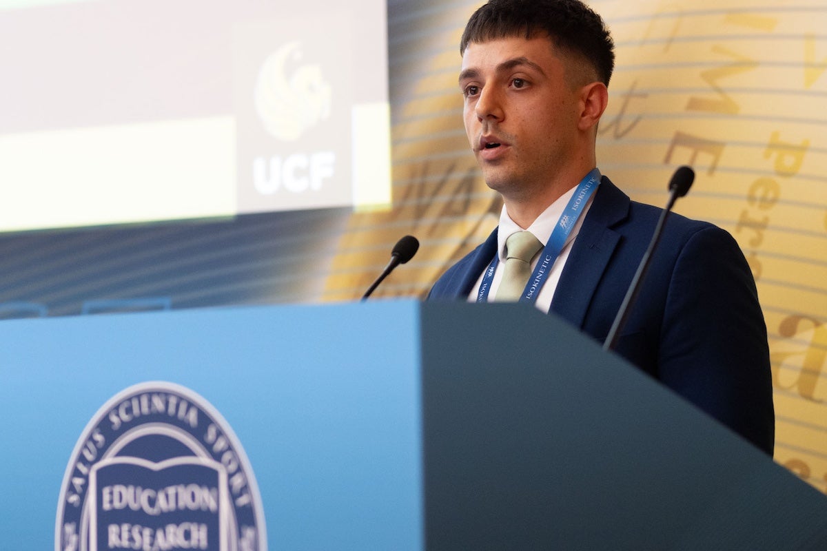 UCF alumnus Jeremy Wydra speaks at a conference podium with a UCF logo displayed on the screen behind him.
