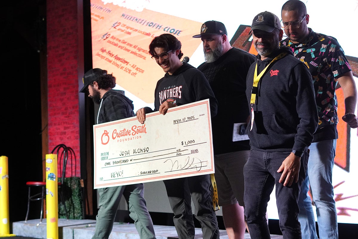 Man holds life size check on stage surrounded by a group of people. 