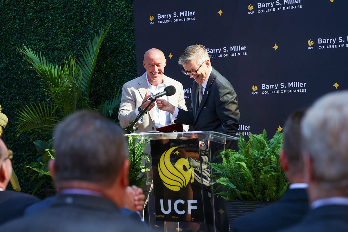 Paul Jarley stands at UCF podium in front of a seated crowd, smiling while looking down, as he hands a key to Barry Miller standing next to him.