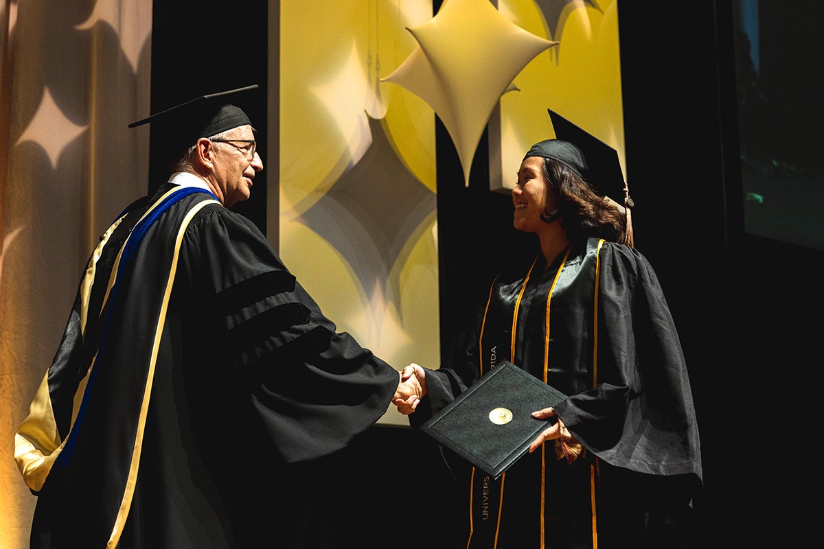 College of Business Dean Paul Jarley, wearing traditional graduation attire, shakes the hand of young Asian graduate holding a black square folder on stage with gold diamond stars handing in the background.