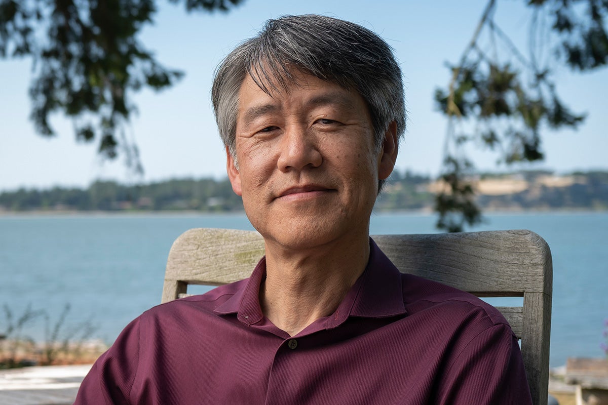 Portrait of Asian man in maroon polo shirt, seated in chair under shade of tree with water in the background.