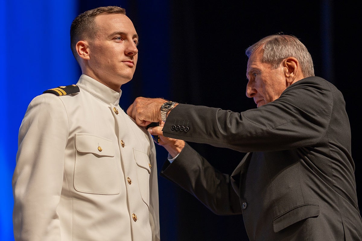 Older man wearing black suit jacket pins medal on left shoulder of young man in dress military uniform