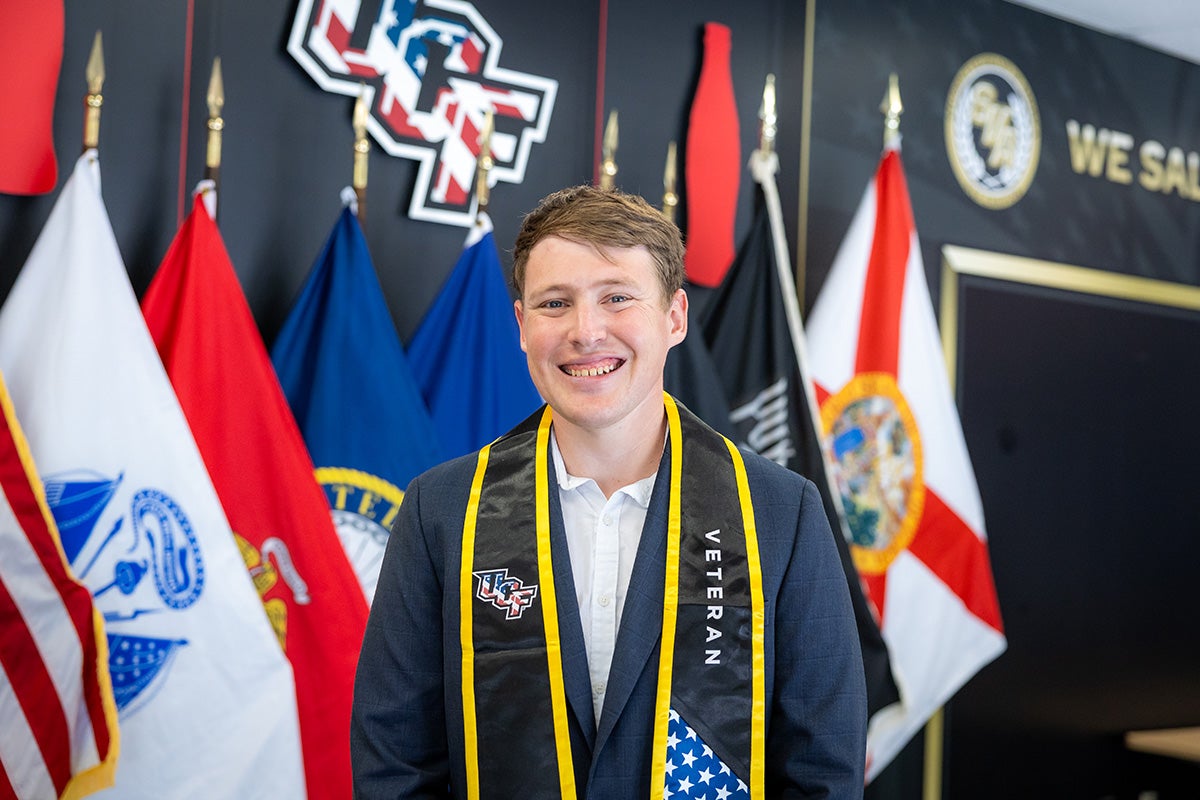 College male with sandy brown hair wearing blue jacket, white dress shirt and black and gold stole with UCF logo stands in front of military flags and wall with UCF -American flag logo 