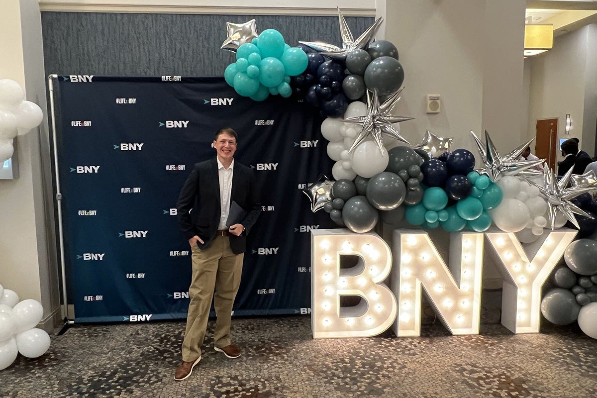 College male with sandy brown hair wearing dark suit jacket, white dress shirt and khaki pants stands in front of BNY media backdrop with large BNY light up mylar letters and balloons next to him.