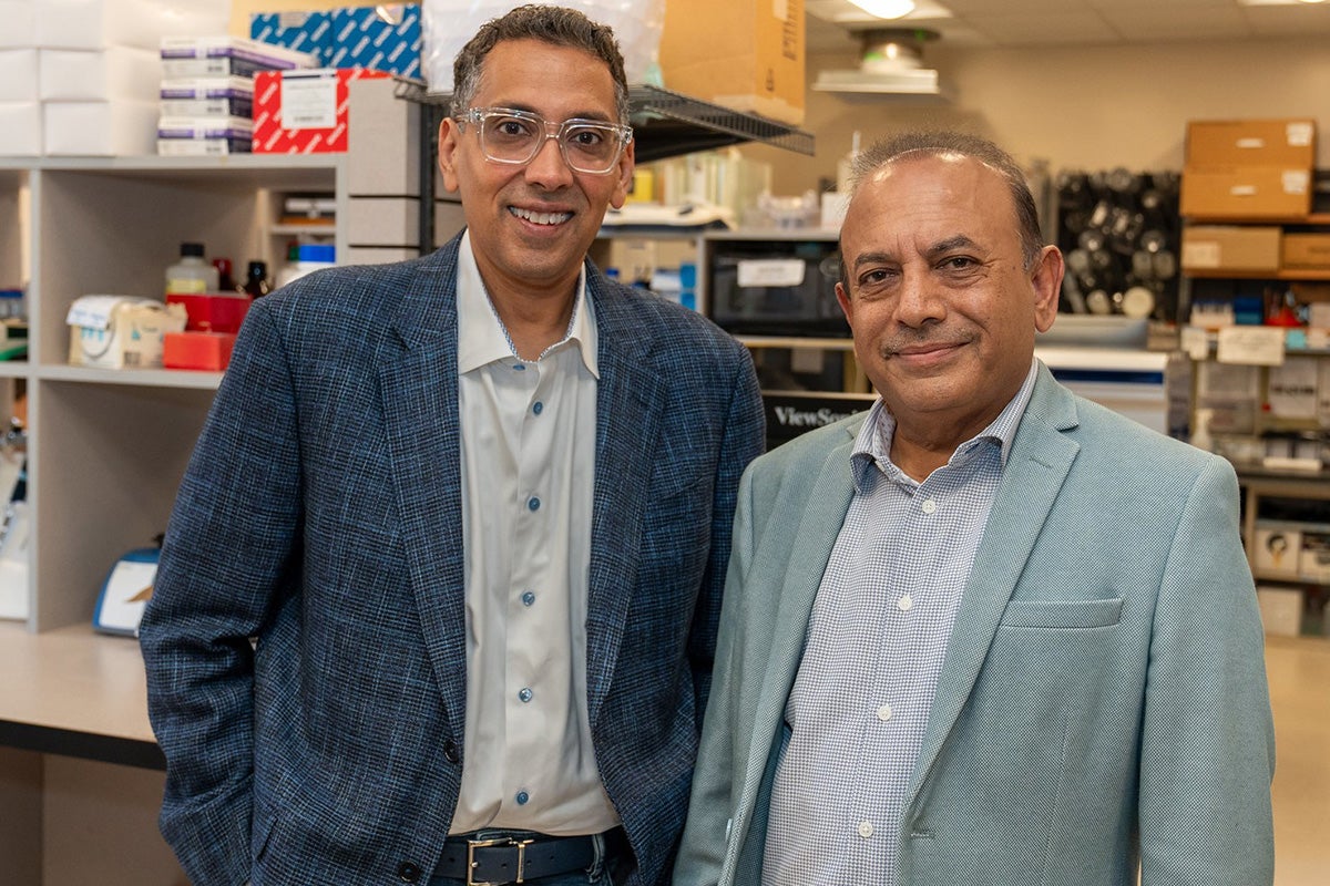 Two men in business jackets pose next to each other in front of shelving with stacks of materials in lab setting