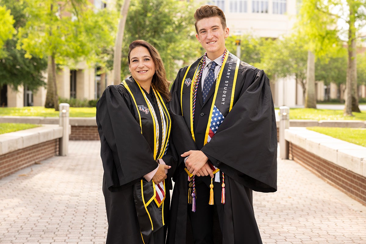 Brunette woman wearing black and gold graduation gown stands next to taller young man wearing black and gold graduation gown on brick patio with trees in background