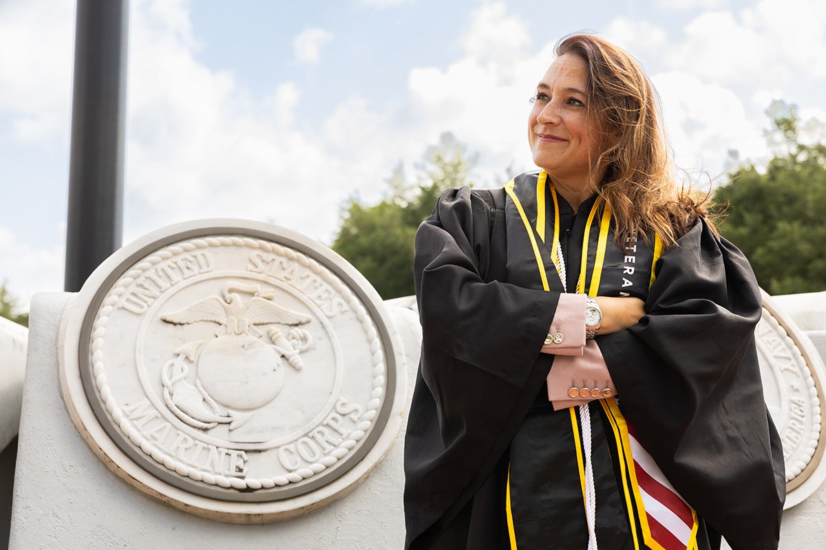 Brunette woman wearing black graduation gown and red white and blue military stole stands with arms crossed in front of concrete statue of seal of US Marines. 