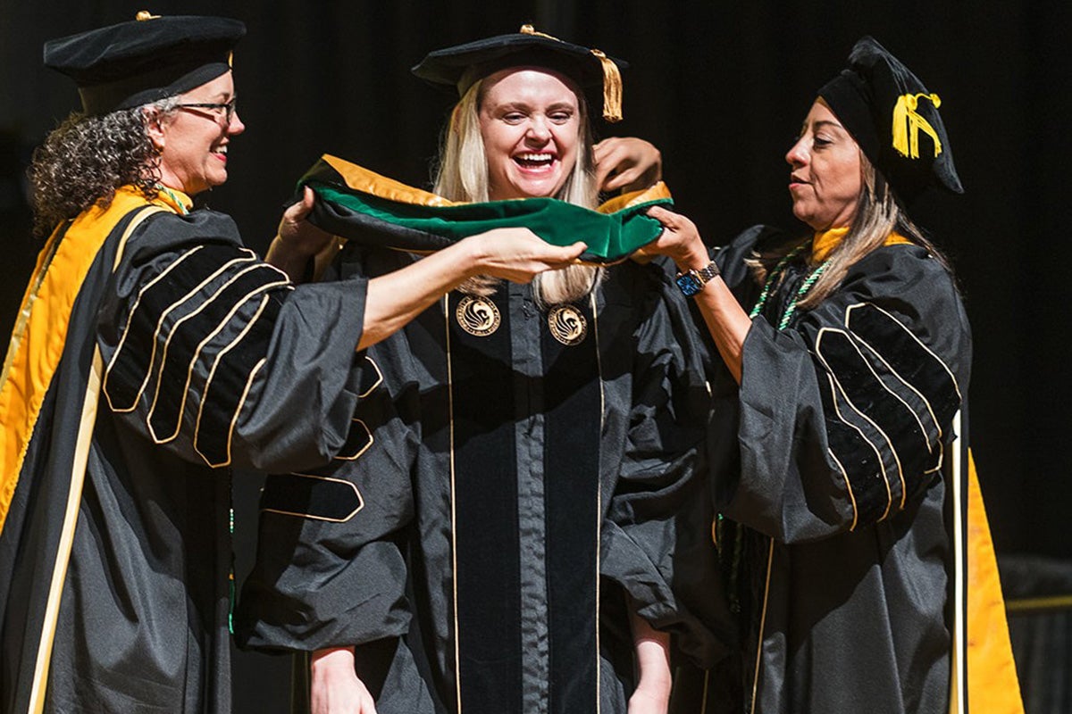Blonde woman is flanked by two older women, all wearing black graduation gowns, as they place gold and green hood over center woman's shoulders