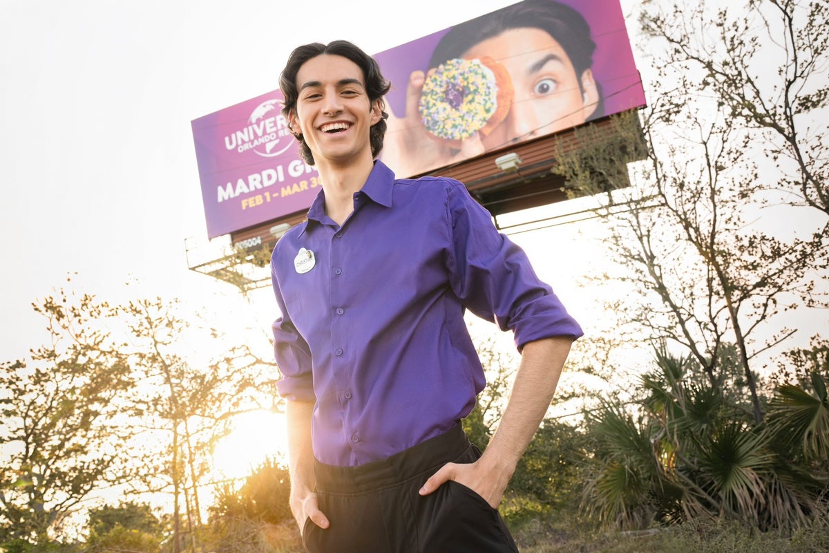 Christian Spencer wearing a Universal Orlando Resort uniform while he stands in front of a Universal Orlando Resort billboard he's featured on