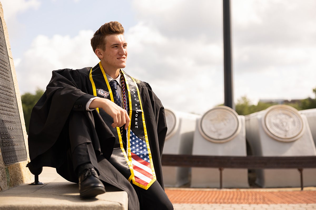 Young man wearing black graduation gown with military red white and blue stole sits in front of veterans concrete memorial outside.