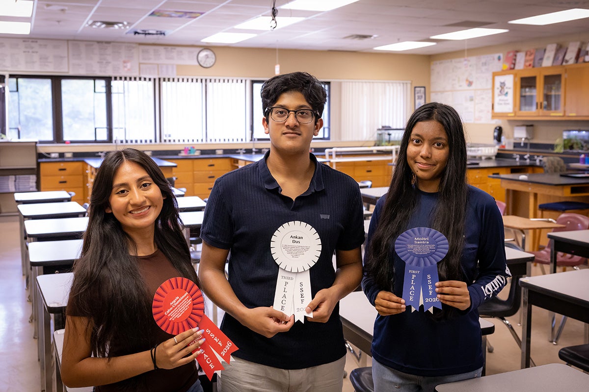 Three high school students posing in classroom with rows of desk and windows in background. Shorter brunette young woman on left holds red ribbon, middle taller young man in center holds white ribbon, young brunette woman on right holds blue ribbon.