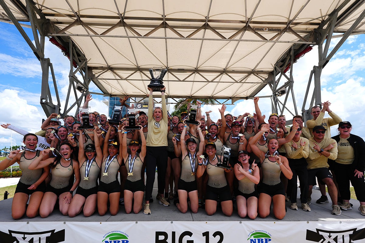 UCF rowing team in gold tops and black shorts gathers on stage under white awning with Mara Allen in center holding up Big 12 trophy on partly cloudy day