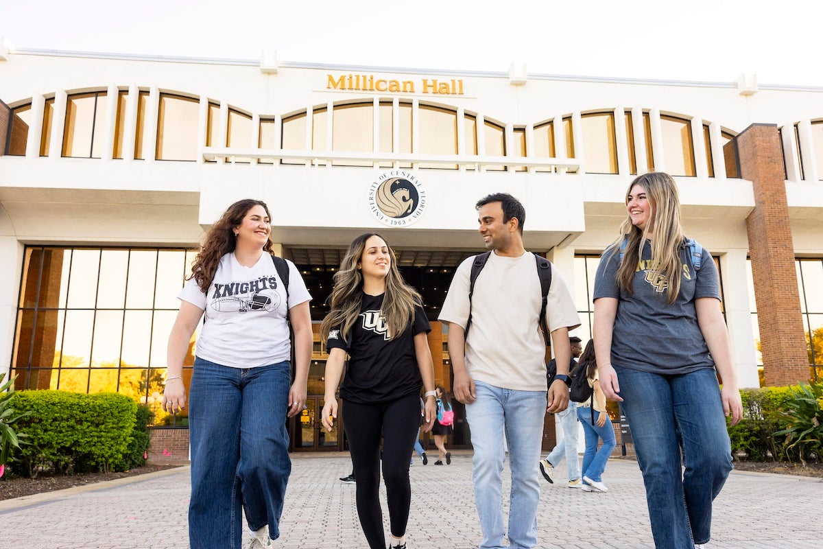 Four students walk together outside Millican Hall on UCF's main campus, smiling and talking.