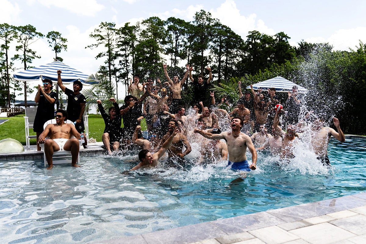 Members of UCF baseball team splash in pool with trees behind them.