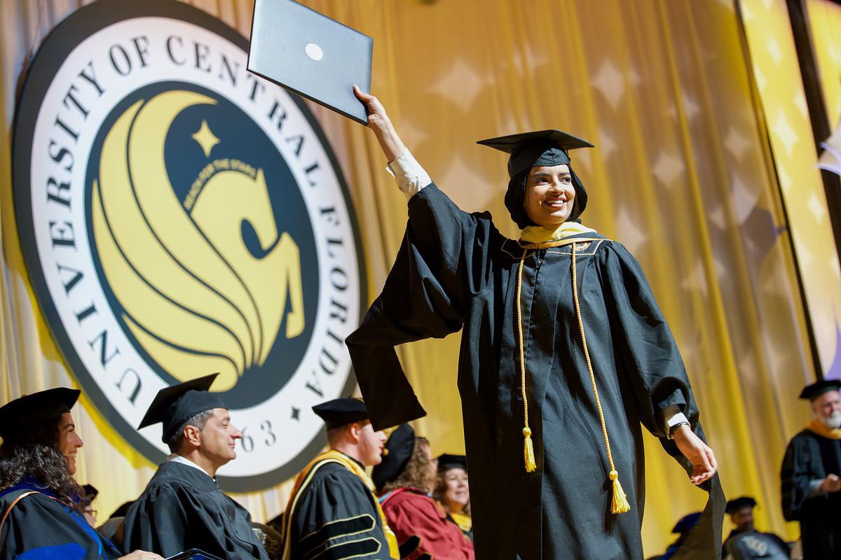 Woman in black cap and gown with gold tassel holds diploma folder in right hand high as she crosses stage with UCF Pegasus seal in background on gold curtains