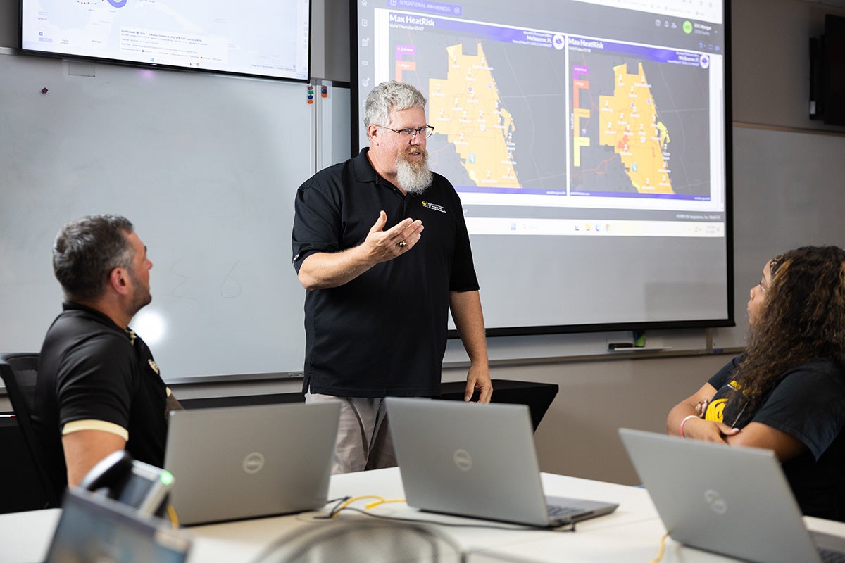 Man with gray hair and beard stands in front of screen with weather maps of Florida projected, talking to two seated individuals at desks with gray Dell laptops opened