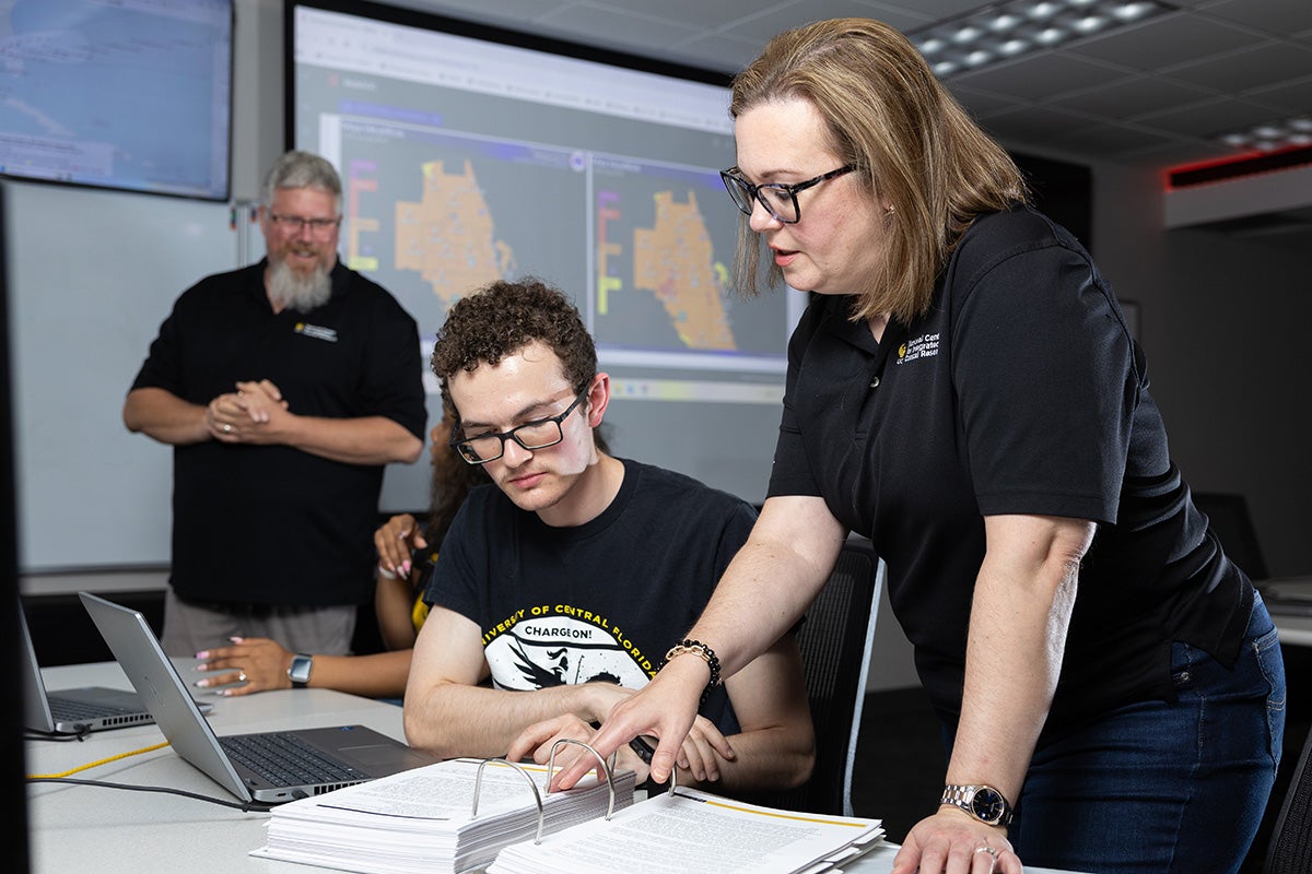 Woman with shoulder length hair and glasses wearing black polo shirt hovers next to desk and man with glasses seated as she points out something in a binder full of papers.