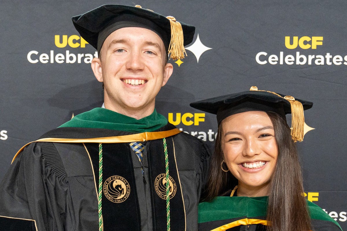 Paxton Threatt and Isabella Castellano, wearing black graduation robes with green trim and black caps with gold tassels, pose in front of back drop with words that read UCF Celebrates.