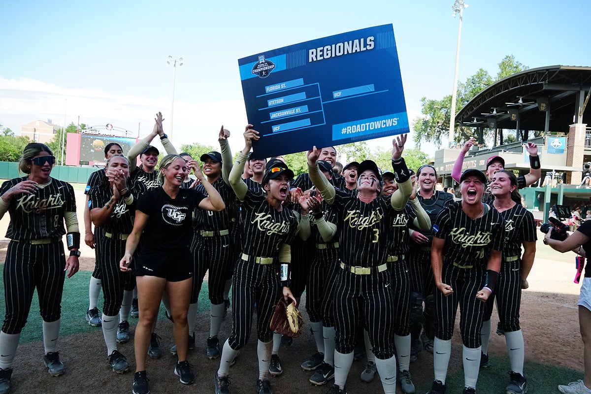 UCF softball team, wearing black pin stripped uniforms, holds blue NCAA Regional bracket poster in air as they celebrate on the field.