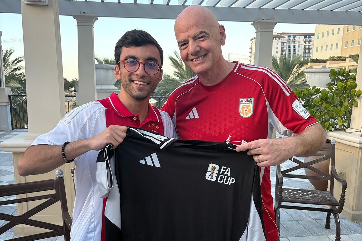 Dark haired young man wearing glasses and white shirt holds black FIFA uniform while standing next to FIFA president Gianni Infantino in red shirt