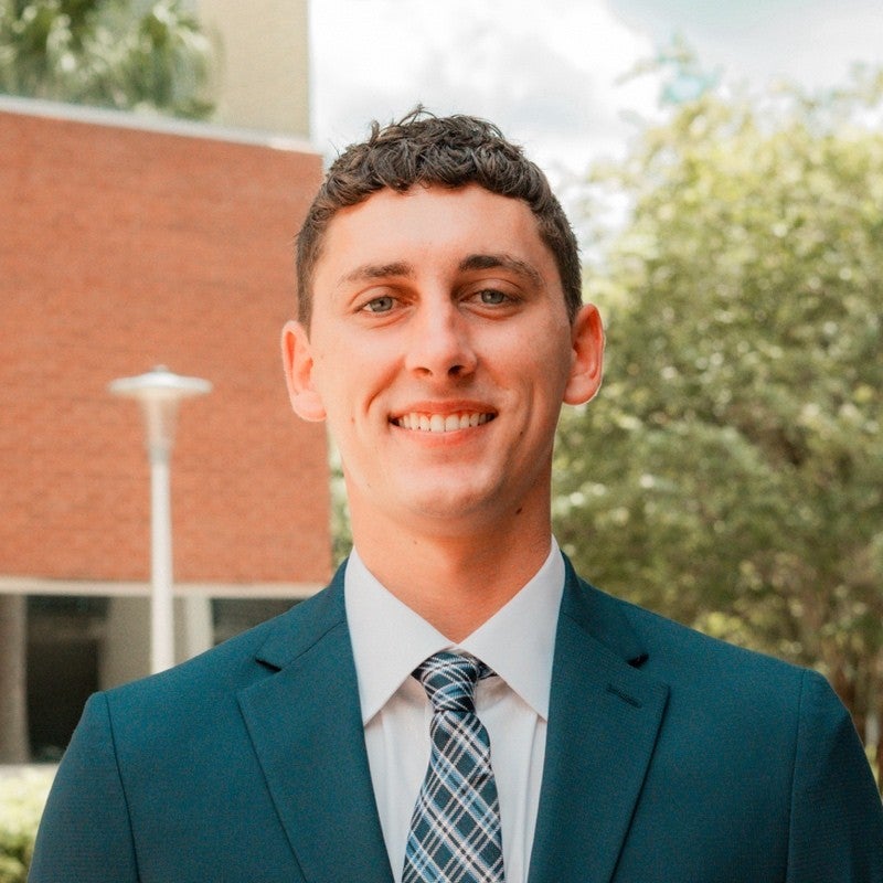 Portrait of smiling man in blue business jacket and plaid tie standing outside with green tree and brick building in background