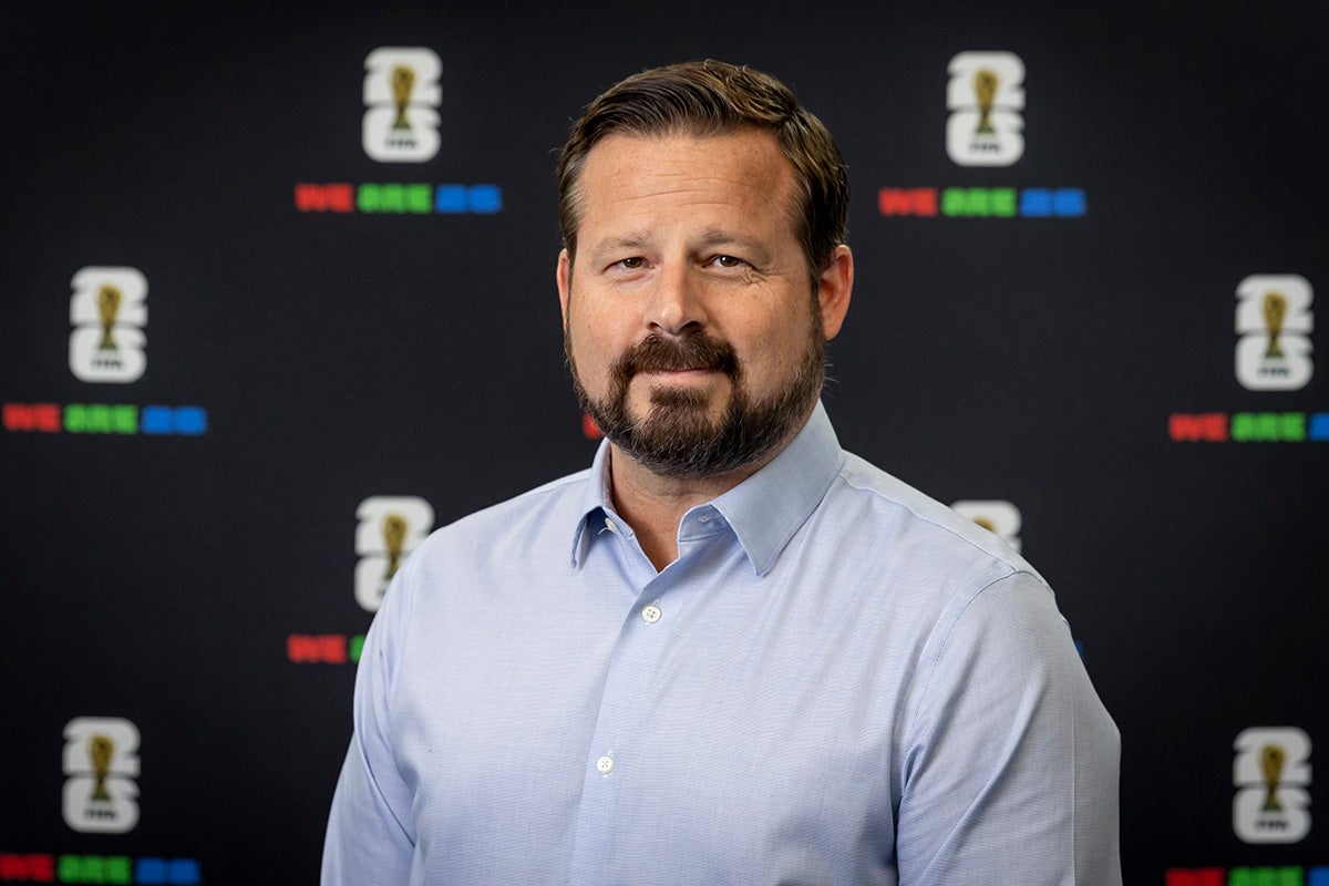 Bearded man with brown hair wearing light blue long sleeve collar shirt stands in front of FIFA backdrop