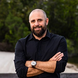 A man wearing a black rolled-sleeve shirt stands with his arms folded and smiling.