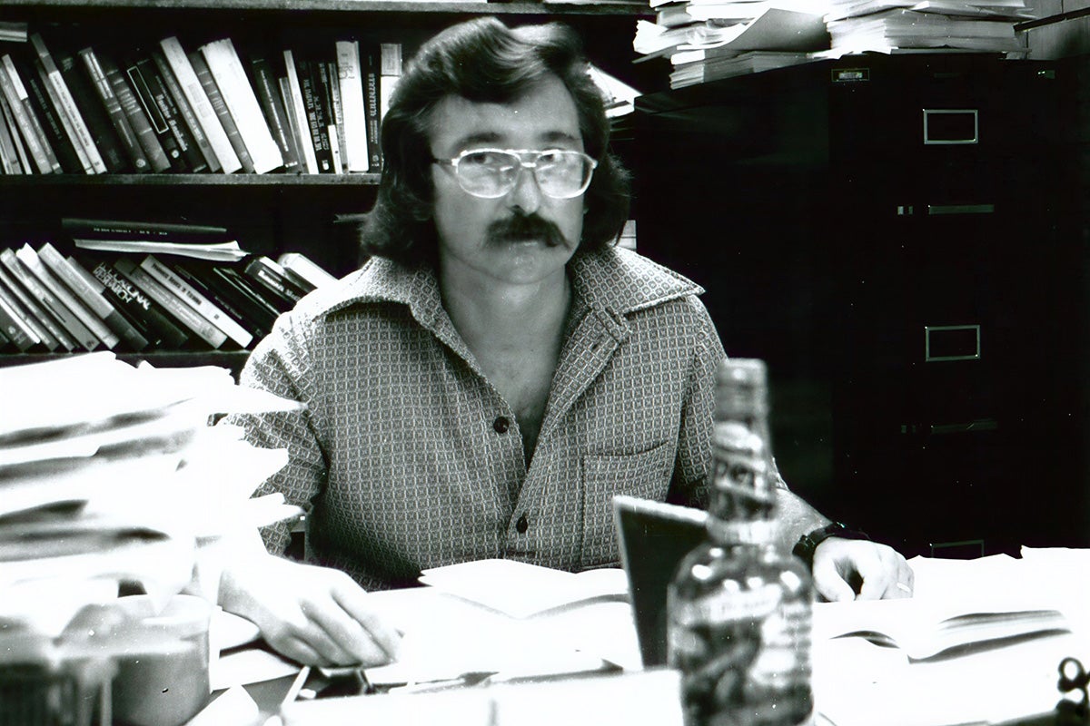black and white photo of dark-haired mustached man with 1970s style classes, long sleeve button shirt, sitting at a desk surrounded by stacks of papers.
