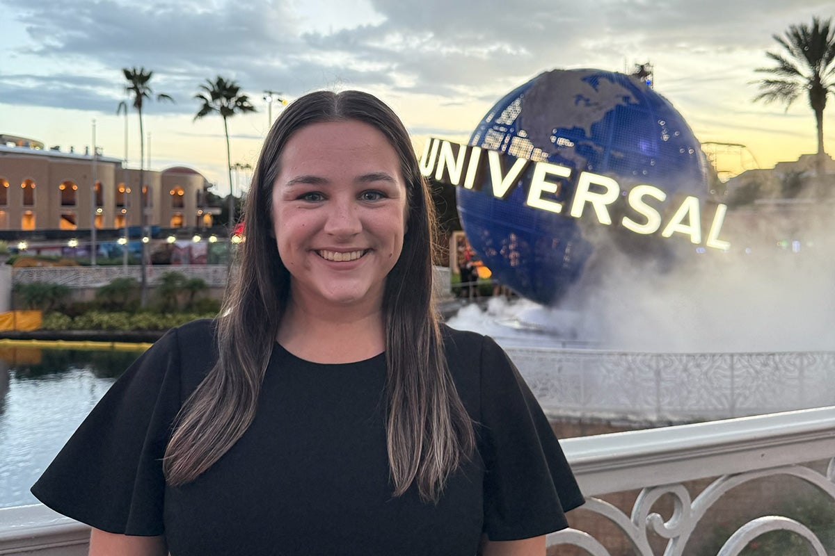 Brunette woman in black blouse stands in front of iconic Universal globe sign at Universal Orlando Resort as sun sets
