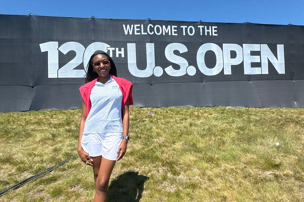 Woman in white outfit with red sweater draped over shoulders poses in front of a sign that reads Welcome to the 126th U.S. Open