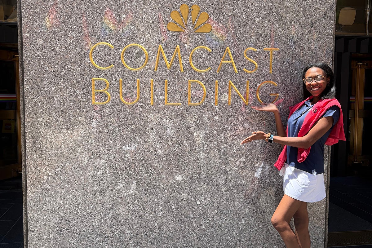 Woman wearing red sweater draped around her shoulders with blue top and white skirts poses in front of gray slate building with the NBC logo and Comcast Building etched in gold lettering