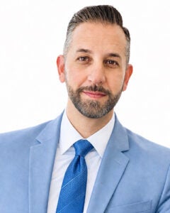Professional head shot of bearded man in light blue jacket, white collar shirt and dark blue tie