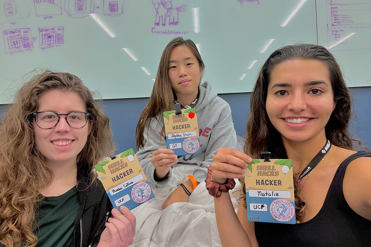 Three women hold up lanyards with credentials that read Shell Hacks Hackers while sitting in front of a whiteboard in classroom setting