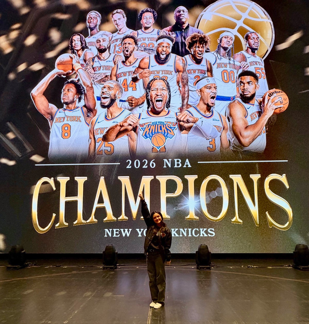 Woman in long sleeve shirt and pants stands in front of graphic of New York Knicks team with the worlds 2026 NBA Champions projected behind her