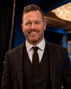 Professional head shot of bearded man with dark suit and tie in room with crystal chandelier 