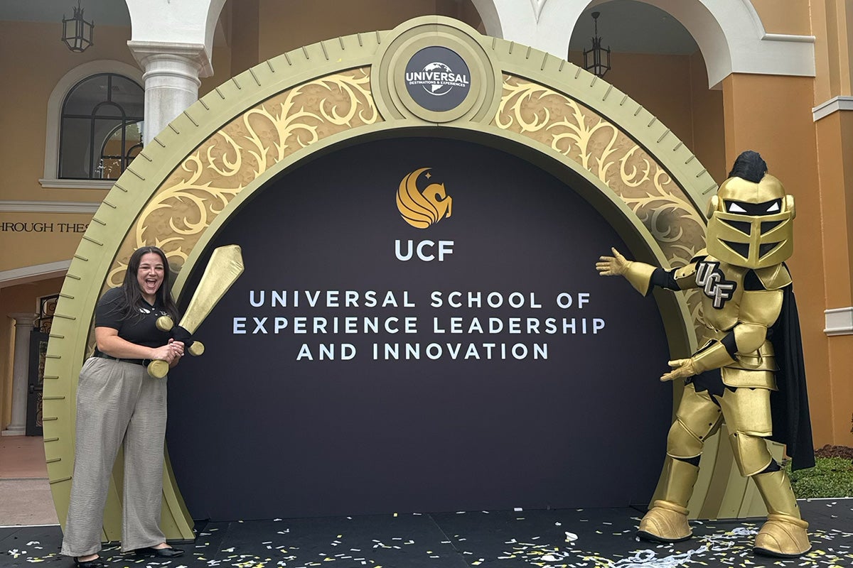 Brunette woman in black top and gray pants holds mascot sword on stage with Knightro flanking the other side of a gold semi-circle sign that reads UCF Universal School of Experience Leadership & Innovation 