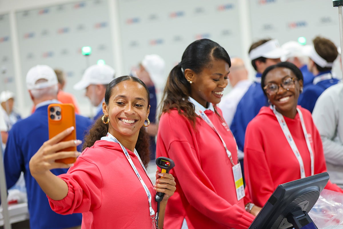 Three Black women wearing red sweaters pose for a selfie while working at a table with a scanner and monitor