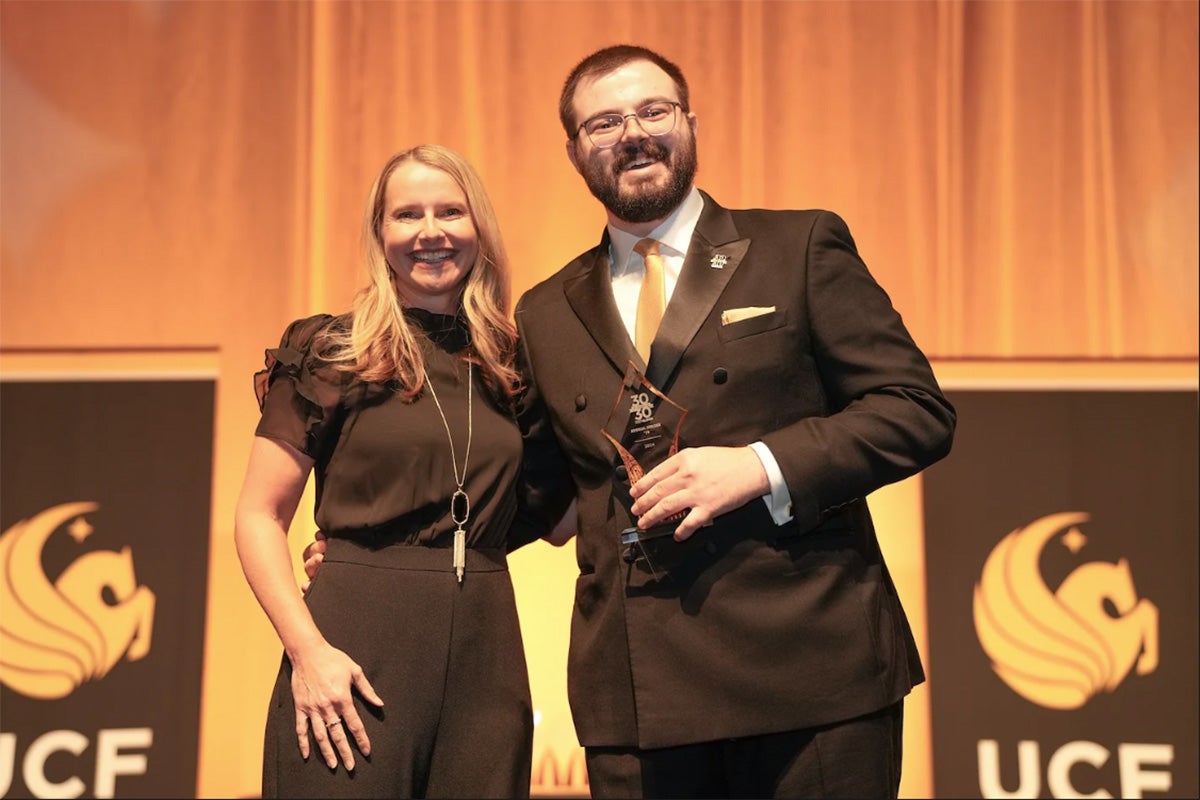 Blonde woman in black jump suit links arms with man in suit holding glass award on stage with UCF banners behind them
