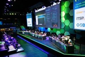 Teams compete at computer stations during the 2019 Wicked6 cybersecurity tournament, with large screens displaying the competition and sponsor logos.